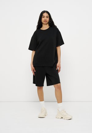 Femeie cu păr lung și închis la culoare, purtând un tricou negru supradimensionat, pantaloni scurți negri până la genunchi, șosete albe și adidași albi masivi, stând în fața unui fundal simplu.