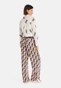 Transparente weiße Langarmbluse mit Ananas-Muster und rosa weiten Hosen mit einem ähnlichen Ananas-Design. Flache Sandalen.