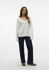 Pull-over blanc à manches longues et col en V avec des fentes sur les côtés, associé à un jean bleu foncé et des baskets marron sur un fond uni.