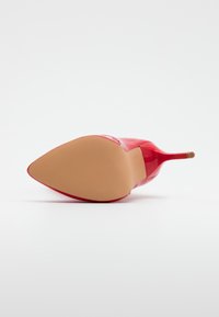 Czerwona lakierowana skórzana szpilka leżąca na boku, ukazująca spiczasty obcas i smukły obcas na białym tle.