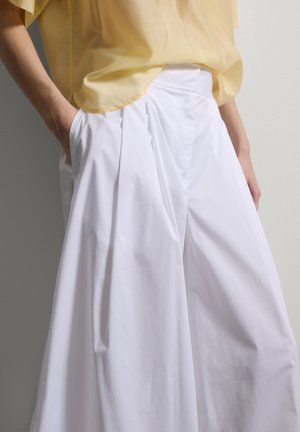 Persona con pantalones blancos de pierna ancha con pliegues y una blusa amarilla holgada, con las manos metidas en los bolsillos del pantalón.