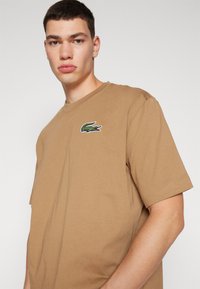 Giovane uomo che indossa una maglietta corta marrone con un logo di coccodrillo verde sul petto, che guarda direttamente verso la fotocamera.