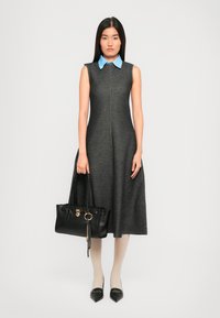 Woman wearing a sleeveless dark gray midi dress with a blue collar, holding a black handbag, paired with white tights and black pointed shoes.