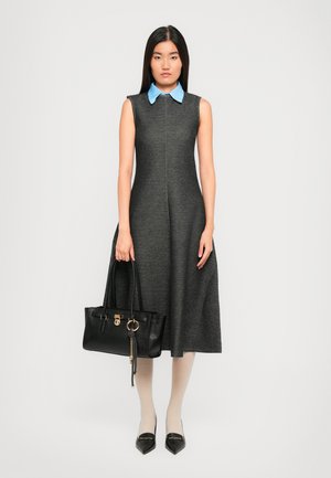 Femme portant une robe midi sans manches gris foncé avec un col bleu, tenant un sac à main noir, assortie avec des collants blancs et des chaussures à talons noires.