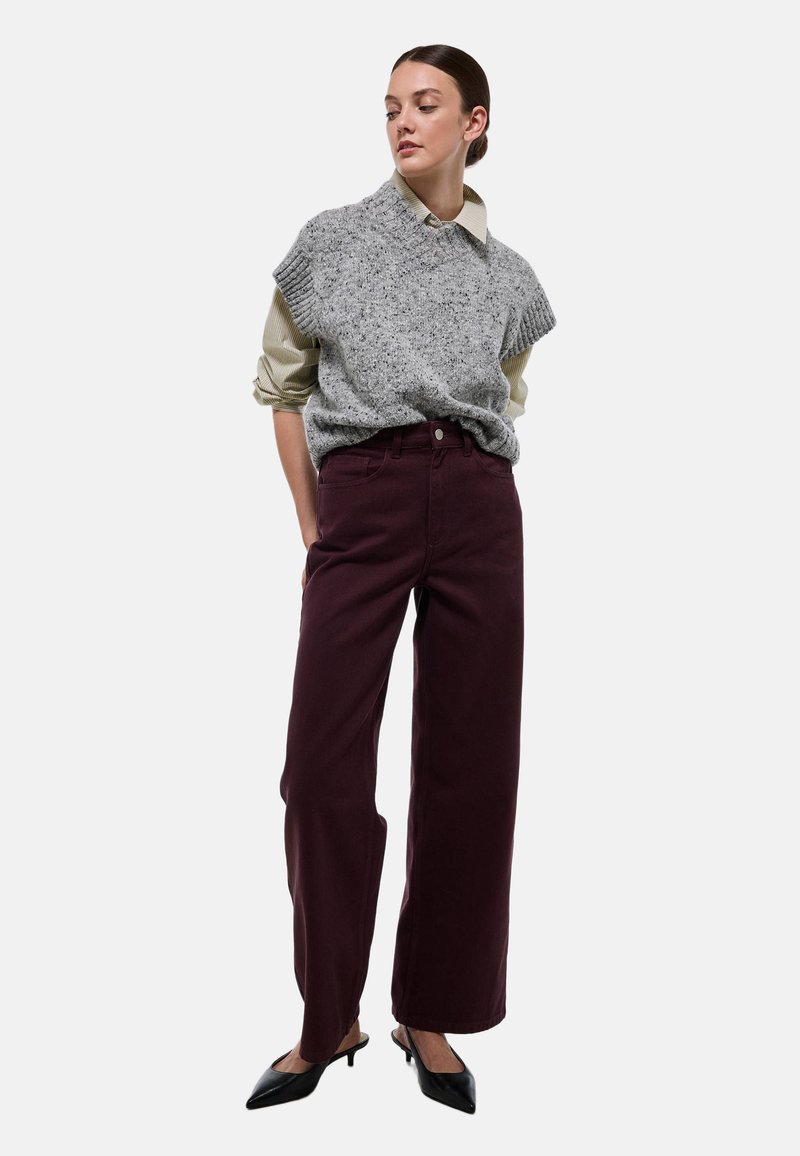 Un pull en tricot gris sans manches porté par-dessus une chemise à col rayé, associé à un pantalon large bourgogne et des escarpins noirs à bout pointu.