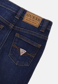 Mörkblå denimjeans med en läderetikett präglad med "GUESS" och en triangulär lapp på bakfickan med kontrasterande sömnad.