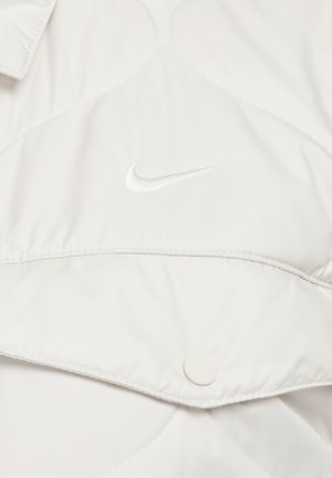 Šviesiai pilkas siuvinėtas švarkas su mygtukų uždarymu ir išsiuvinėtu baltu Nike logotipu. Lygus paviršius su šiek tiek blizgiu finišu.