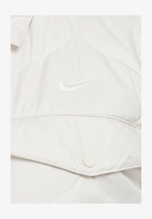 Šviesiai pilkas siuvinėtas švarkas su mygtukų uždarymu ir išsiuvinėtu baltu Nike logotipu. Lygus paviršius su šiek tiek blizgiu finišu.