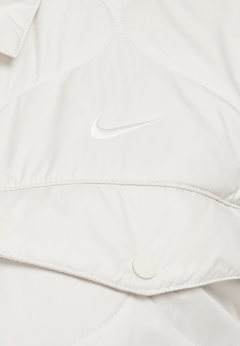 Veste matelassée gris clair avec fermeture à boutons et logo Nike brodé en blanc. Texture lisse avec une finition légèrement brillante.