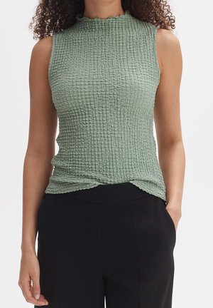 Sleeveless sage green textured top with ruffled mock neck, worn with black pants, model standing with one hand in pocket.