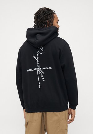 Man with dreadlocks wearing a black hoodie with "WORLDWIDE STANDARD" and vertical "Only Love" text, paired with beige cargo pants.