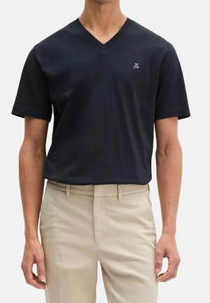 Mann trägt ein schlichtes schwarzes T-Shirt mit V-Ausschnitt und kurzen Ärmeln sowie beige Hosen und steht mit entspannten Armen seitlich.