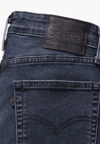 Temno modre jeans hlače iz denima z usnjenim krojem na pasu, ki ima embosirano besedilo in grafični dizajn. Vključujejo zadnji žep s podrobnostmi v šivanju.