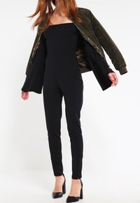 Svart jumpsuit med axelbandslös urringning, figursydd siluett, kombinerad med en olivgrön bomberjacka. Modellen bär svarta högklackade skor.