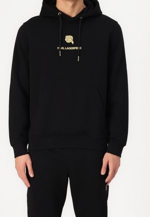 Svart hoodie i mjukt tyg, med en central golden logotyp och dragsko i huvan. Inkluderar en framficka och ribbade muddar.