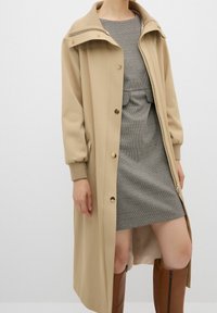 Abrigo beige con cuello alto, botones dorados y puños elásticos, llevado sobre un vestido gris a cuadros, combinado con botas marrones hasta la rodilla.