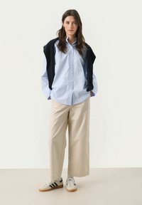 Light blue and white striped button-up shirt with a dark sweater draped over shoulders, paired with beige wide-leg pants and gray sneakers.