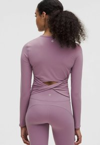 Top athletic cu mâneci lungi dintr-un material ribbed de culoare mov deschis, având un design cu spate răsucit și detalii cu decupaj, cu o croială ergonomică.