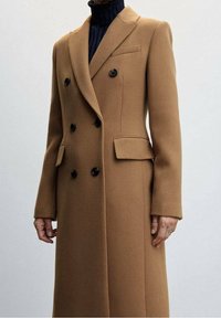 Cappotto in misto cammello doppio petto con bottoni neri, rever a punta, tasche laterali e silhouette strutturata, indossato da un modello.