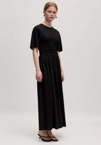 Zwarte maxi-jurk met korte mouwen, gerimpelde bovenkant en een zwierige rok. Gelaagd met zwarte platte sandalen, die een soepele stoftextuur tonen.