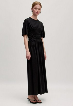 Zwarte maxi-jurk met korte mouwen, gerimpelde bovenkant en een zwierige rok. Gelaagd met zwarte platte sandalen, die een soepele stoftextuur tonen.