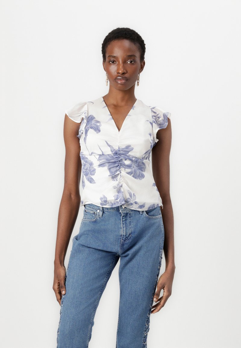 Blusa floreale bianca con motivi blu, scollatura a V e maniche corte a volant, abbinata a jeans di denim azzurro chiaro con decorazioni laterali.