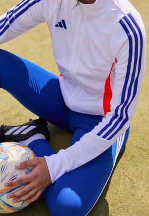 Henkilö pukeutuneena valkoiseen ja siniseen Adidas-verryttelypukuun istuu ruohikolla, pitäen värikästä jalkapalloa yhdellä kädellä.