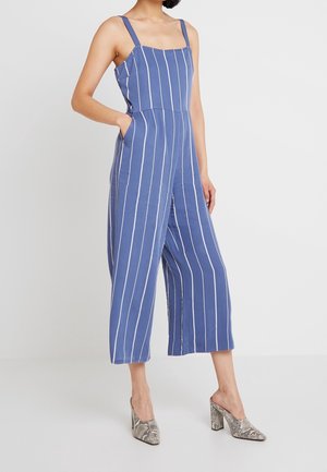 Frau trägt einen blauen Jumpsuit mit weißen vertikalen Streifen und weitem Bein, kombiniert mit hochhackigen Schuhen im Schlangenledermuster.