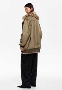 Chaqueta bomber verde oliva con ribete de pelo sintético, puños y dobladillo acanalados. Corte holgado combinado con pantalones anchos negros. Textura de tela suave y brillante.
