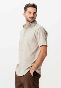 Mand med kort hår og skæg iført lys beige kortærmet skjorte med knapper og brune bukser, hænderne i lommerne, kigger til siden.