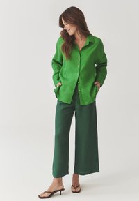 Groene linnen button-up shirt met een ontspannen pasvorm, gecombineerd met donkergroene wijde broek en zwarte sandalen. Gladde textuur, minimale accenten.
