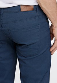 Marineblaue, strukturierte Hose mit einem Taillenlabel, ausgestattet mit Gesäßtaschen und geradem Schnitt. Detaillierte Nähte entlang der Säume.