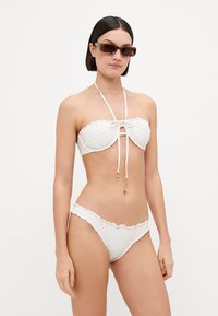 Beli bikini komplet s teksturiranim vzorcem, ki vključuje bandeau zgornji del s pentljo in nastavljivimi trakovi, v kombinaciji z nizkimi hlačkami.