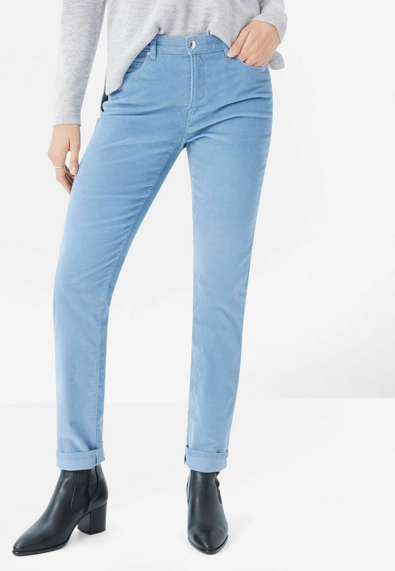Pantalon bleu clair à jambes droites en coton à la texture douce, doté d'une fermeture boutonnée à l'avant et d'ourlets retroussés, porté avec des bottines noires.
