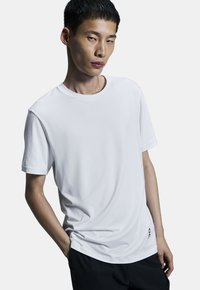 White short-sleeve T-shirt made of smooth fabric, featuring a round neckline and a subtle logo on the bottom left hem.