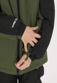 Grön och svart vattentät jacka med elastiska mudd, dragkedjestängning och justerbar midja. Har logotypdetalj på ärmen.
