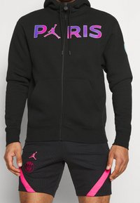 Sudadera negra con cremallera y la palabra "PARÍS" en texto degradado de púrpura a rosa, con un bolsillo tipo canguro. Combinada con pantalones cortos negros con acentos rosas.