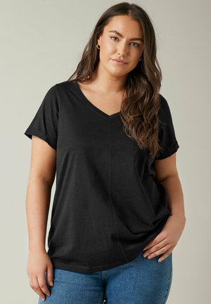 Junge Frau mit langen braunen Haaren, die ein schwarzes V-Ausschnitt-T-Shirt und blaue Jeans trägt, steht mit einer Hand in der Hüfte vor einem neutralen Hintergrund.