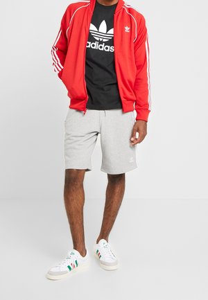 Červená sportovní bunda s bílými pruhy, černé tričko s logem, šedé šortky a vícebarevné tenisky s bílou podrážkou.