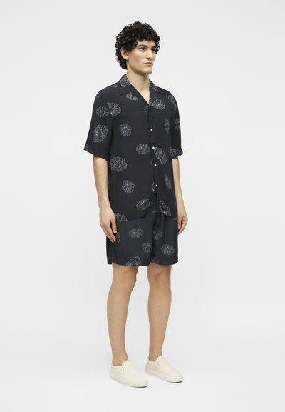 Νέος άνδρας με σγουρά μαλλιά που φοράει μαύρο κοντομάνικο πουκάμισο με κουμπιά και ταιριαστά σορτς με λευκά λουλουδάτα σχέδια και λευκά παπούτσια slip-on.