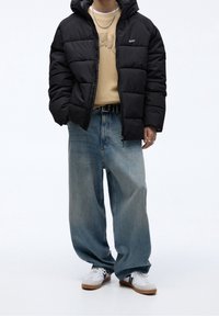 Giacca a vento nera con cappuccio, felpa beige sotto, jeans blu chiaro larghi e sneakers bianche con strisce nere e suole marrone.