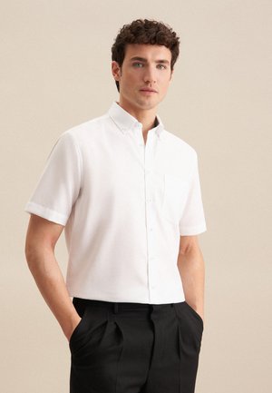 Chemise blanche à manches courtes avec col boutonné, poche poitrine unique et texture lisse. Associée à un pantalon noir.