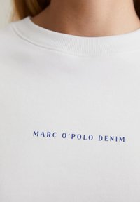 Witte sweatshirt met geribbelde halslijn, met blauwe tekst "MARC O'POLO DENIM" centraal op de voorkant gedrukt. Gladde katoenstructuur.