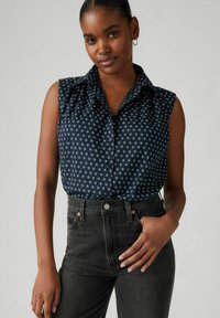 Blusa senza maniche blu navy con motivo floreale bianco, abbottonatura frontale e dettaglio arricciato sulle spalle, abbinata a jeans scuri a vita alta.