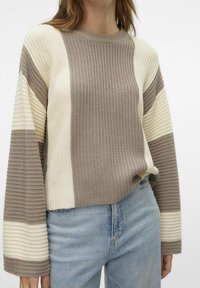 Pull en maille avec rayures horizontales en beige, crème et taupe, présentant un design côtelé texturé et des manches larges. Associé à un jean bleu clair.