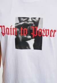 Hvit bomulls-tanktop med et sentralt svart-hvitt fotografi av en hånd som holder en gjenstand, med teksten "Pain to Power" i fet rød gotisk skrift.