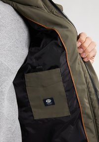 Intérieur d'une veste rembourrée olive avec doublure noire, passepoil orange et une petite poche avant arborant une étiquette de logo.
