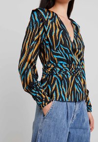 Blusa nera con motivo a righe blu e oro, scollatura a V profonda, spalle arricciate e vita cinchata. Abbinata a pantaloni in denim blu chiaro.