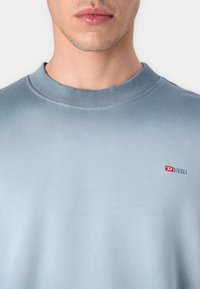Světle modrý crewneck svetr z měkké tkaniny, s žebrovaným límečkem a malým červeným a modrým logem "DIESEL" na hrudi.
