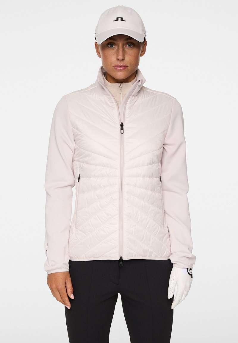 En person står vänd mot kameran, iklädd en ljusrosa jacka och vit keps. De håller en golfhandske i vänster hand.